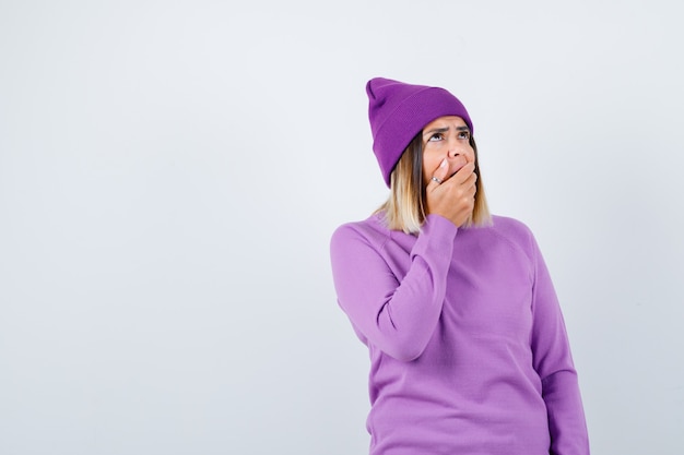 Hübsche Dame im Pullover, Mütze mit Hand auf offenem Mund, nach oben schauend und überrascht, Vorderansicht.