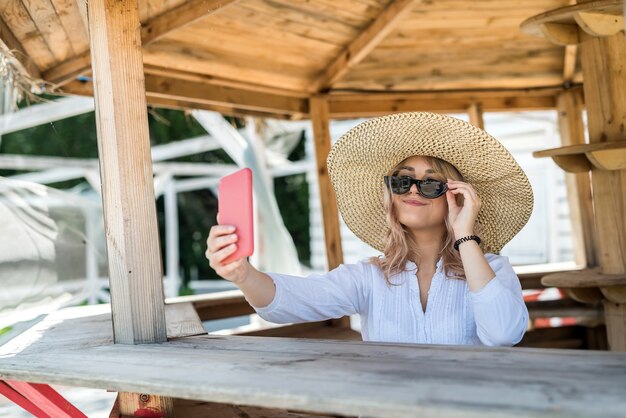 Hübsche Dame mit Zelle und Selfie in der Nähe von Holzpavillon am heißen Sommertag. Entspannen und genießen Sie die Natur