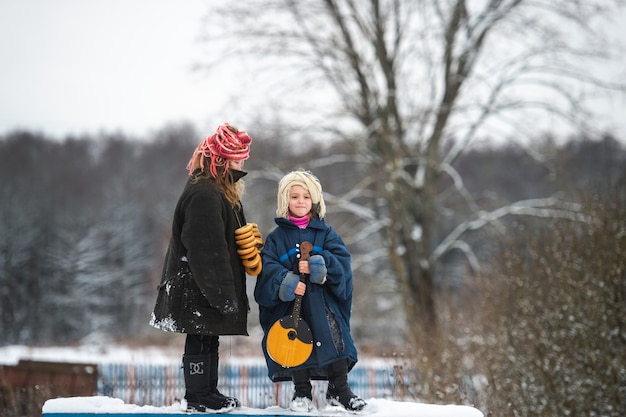 Hübsche Dorfschwestern stehen mit Balalaika und getrockneten ringförmigen Brötchen auf der Bank und lächeln im Winter