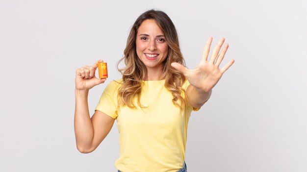 Hübsche dünne Frau, die lächelt und freundlich aussieht, Nummer fünf zeigt und Batterien hält