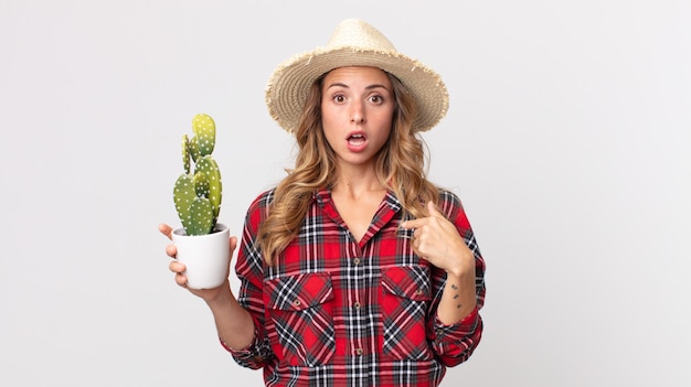 Hübsche dünne Frau, die schockiert und überrascht mit weit geöffnetem Mund aussieht und auf sich selbst zeigt, der einen Kaktus hält. Bauernkonzept