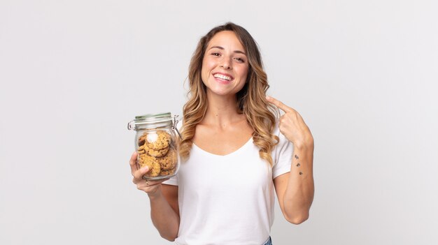 Hübsche dünne Frau, die selbstbewusst auf ihr eigenes breites Lächeln lächelt und eine Glasflasche mit Keksen hält