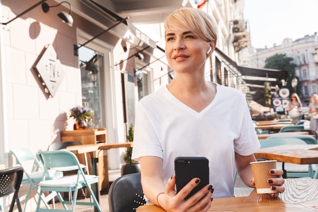 hübsche erwachsene Frau, die weißes T-Shirt mit Handy und kabellosem Kopfhörer trägt, während im Sommer draußen im Café sitzt und Kaffee aus Pappbecher trinkt