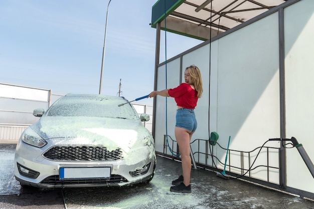 Hübsche Fahrerfrau, die ihr Auto mit Hochdruckschlauch von Schmutz säubert, trägt einen Reiniger auf das Auto auf