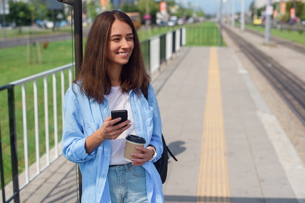 Hübsche Frau benutzt Smartphone und trinkt Morgenkaffee, während sie auf der öffentlichen Station auf Straßenbahn oder Bus wartet.