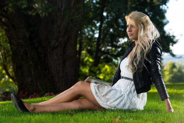 Hübsche Frau, die auf dem Gras mit digitalem Tablet sitzt