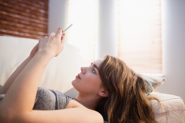 Hübsche Frau, die auf der Couch unter Verwendung des Telefons im Wohnzimmer liegt