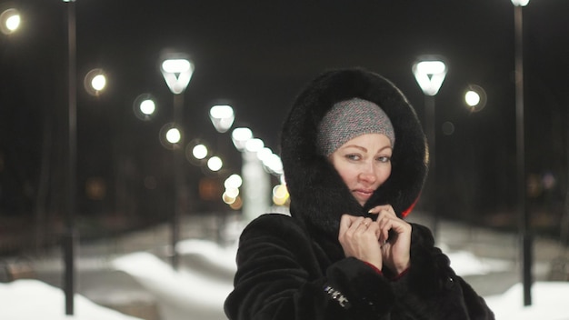 Hübsche Frau, die den schwarzen Pelzmantel entlang geht Outdoor Girl geht in einer Winternacht zwischen den Straßenlaternen der Stadt Snow Park mit Girlanden Schönes sinnliches glückliches Lächeln