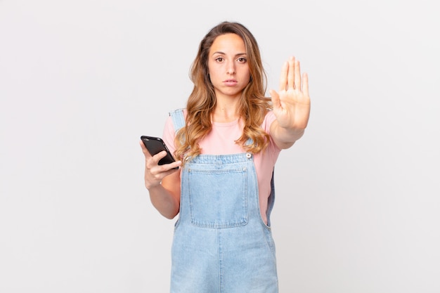 Hübsche Frau, die ernst aussieht und eine offene Handfläche zeigt, die eine Stopp-Geste macht und ein Smartphone hält