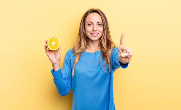 Hübsche Frau, die freundlich lächelt und aussieht und die Nummer eins zeigt, die eine halbe Orange hält