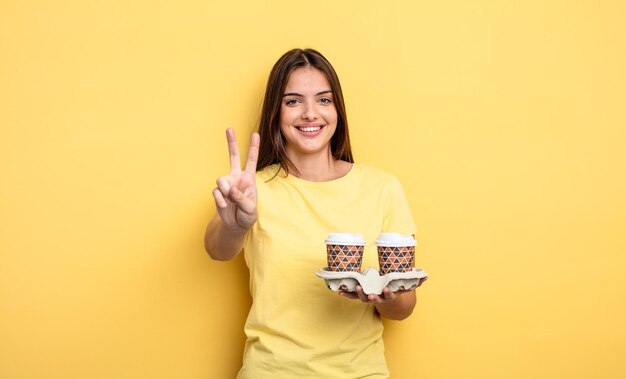 Hübsche Frau, die freundlich lächelt und aussieht und Nummer zwei zeigt. kaffeekonzept zum mitnehmen