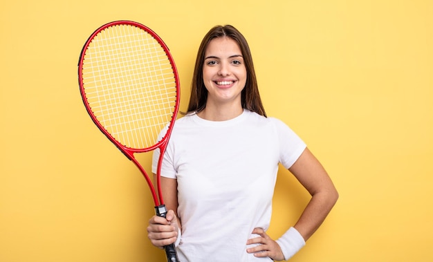 Hübsche Frau, die glücklich mit einer Hand auf Hüfte und überzeugt lächelt. Tennisspieler-Konzept