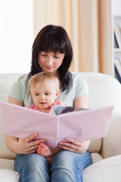 Hübsche Frau, die ihr Baby und ein Buch in ihren Armen beim Sitzen auf einem Sofa hält