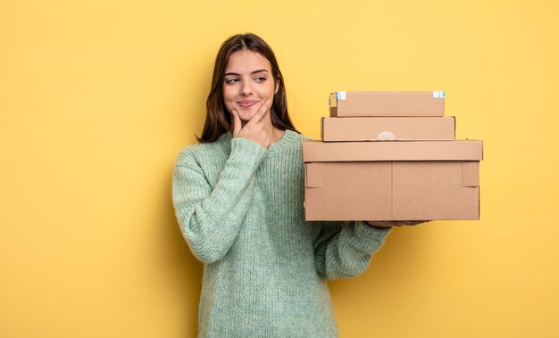 Hübsche Frau, die mit einem glücklichen, selbstbewussten Ausdruck mit der Hand am Kinn lächelt, verpackt Boxen-Konzept