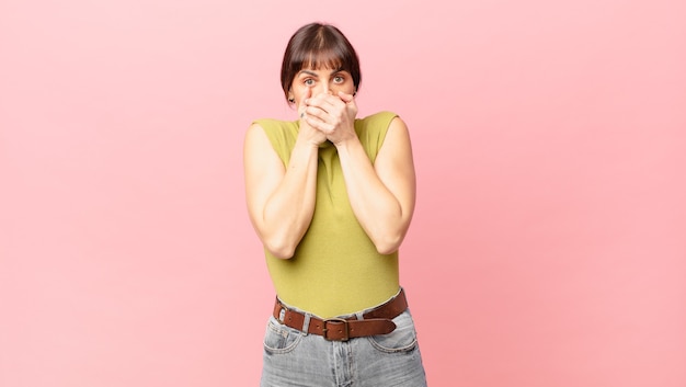 Foto hübsche frau, die mit einem schockierten, überraschten ausdruck den mund mit den händen bedeckt, ein geheimnis hält oder oops sagt