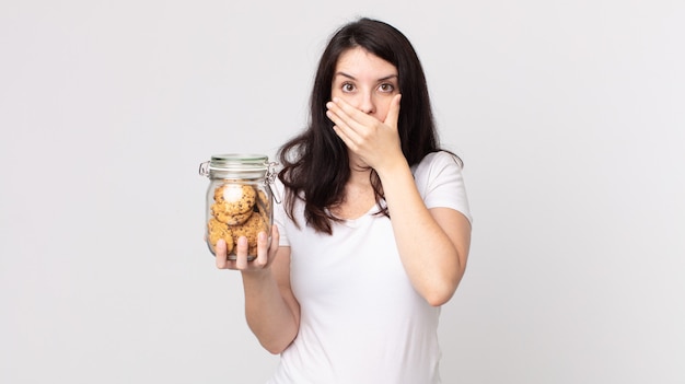 hübsche Frau, die schockiert den Mund mit den Händen bedeckt und eine Glasflasche mit Keksen hält