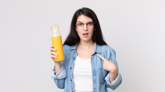 Hübsche Frau, die sich glücklich fühlt und auf sich selbst zeigt, aufgeregt und hält eine Kaffee-Thermoskanne