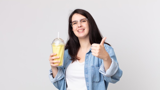 Hübsche Frau, die sich stolz fühlt, positiv mit erhobenen Daumen lächelt und einen Vanille-Milchshake hält