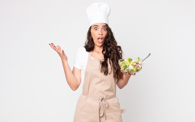 Hübsche Frau erstaunt, schockiert und erstaunt über eine unglaubliche Überraschung, die eine Schürze trägt und einen Salat in der Hand hält