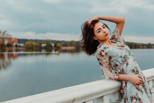 Hübsche Frau gekleidetes blumiges Kleid, das auf dem Pier nahe Flusssee launisches bewölktes Wetter steht