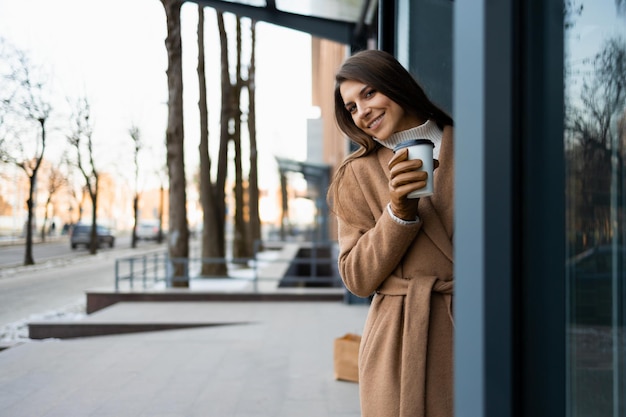 Hübsche Frau im braunen Mantel mit Kaffeetasse im Freien