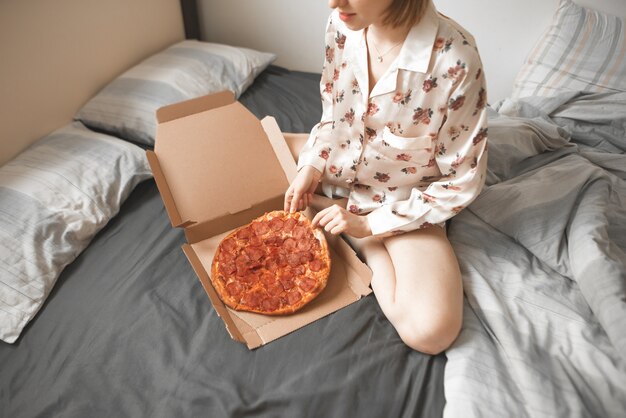 Hübsche Frau im Pyjama sitzt auf einem Bett mit einer Pizzaschachtel und nimmt ein Stück.