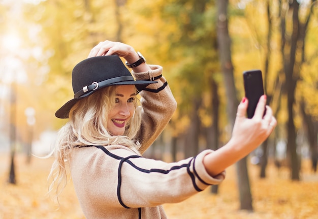 Hübsche Frau im schwarzen Hut, der Selfie mit ihrem Smartphone im Herbstpark macht