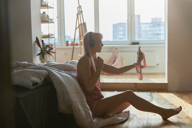 Hübsche Frau im stylischen Pyjama mit Kopfhörern macht Selfie in der Nähe eines großen Bettes auf dem Boden