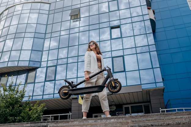 Hübsche Frau im weißen Anzug, die ihren Elektroroller nach der Fahrt hält, während sie auf dem Hintergrund eines modernen Gebäudes steht