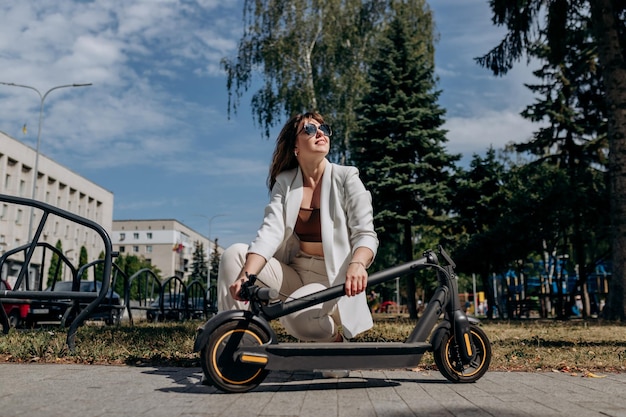 Hübsche Frau im weißen Anzug, die ihren Elektroroller nach der Fahrt zusammenklappt, während sie in der Parklandschaft der Stadt steht