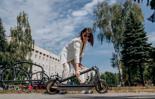 Hübsche Frau im weißen Anzug, die ihren Elektroroller nach der Fahrt zusammenklappt, während sie in der Parklandschaft der Stadt steht