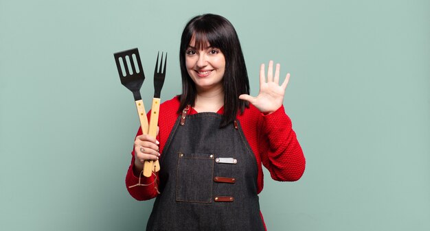 Hübsche Frau in Übergröße, die lächelt und freundlich aussieht, die Nummer fünf oder fünf mit der Hand nach vorne zeigt und herunterzählt