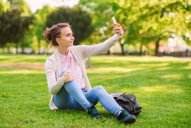 Hübsche Frau in einer rosa Bluse und im blauen Denim, die auf dem Gras sitzen und Selfie tun