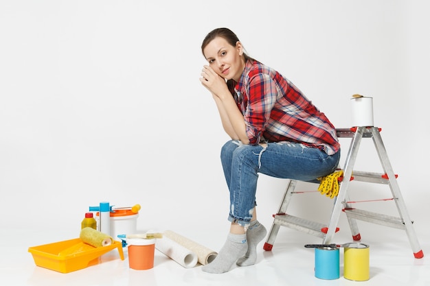 Hübsche Frau in Freizeitkleidung, die auf Leiter mit Instrumenten für Renovierungswohnungsraum lokalisiert auf weißem Hintergrund sitzt. Tapeten, Zubehör zum Kleben, Malwerkzeuge. Hauskonzept reparieren.