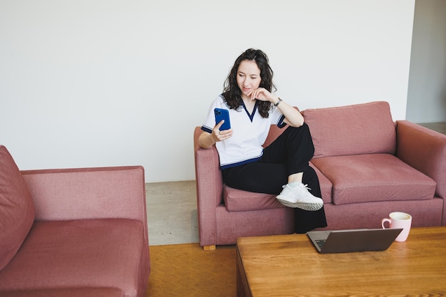 Hübsche Frau in lässiger Kleidung sitzt auf einem bequemen Sofa in einem modernen Café am Tisch mit Laptop und Telefon und arbeitet online Fernarbeit