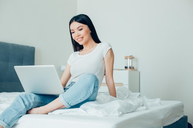 Hübsche Frau lächelt und schaut auf den Bildschirm eines modernen Laptops, während sie damit auf dem Bett sitzt