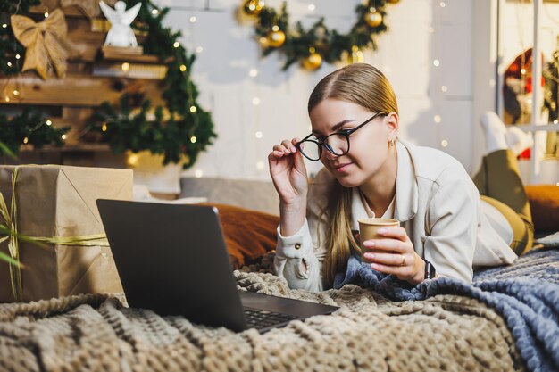 Hübsche Frau mit blonden Haaren in gemütlicher Kleidung mit Kaffee in der Hand mit Laptop, die aus der Ferne arbeitet und auf dem Bett im Zimmer mit Weihnachtsbaum zu Hause lächelt Neujahrsferien