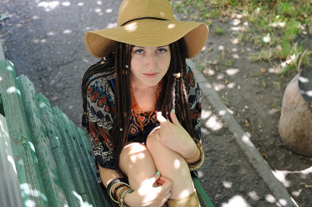 Hübsche Frau mit Dreadlocks, gekleidet in Kleid und Hut im Boho-Stil, Blick in die Kamera, im Freien