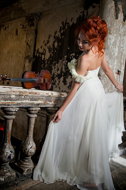 Hübsche Frau mit Geige. Geigerin und Schauspielerin. Mädchen in einem Bild- und Bühnenkleid, Kostüm in einem alten strukturierten Raum hält oder spielt Geige. Konzept des Musikplakats. Autorenraum im Foto