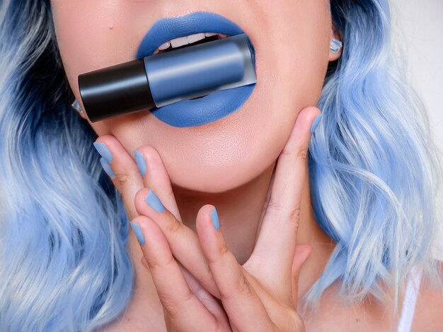 Hübsche Frau mit modischem Lippenstift des Kameraden im Mund, Nahaufnahme. Geerntetes Foto blauer Haardame Lippenstift in den Lippen halten. Make-up-Konzept, Verkauf Kit Idee. Klassische blaue Farbe des Jahres 2020