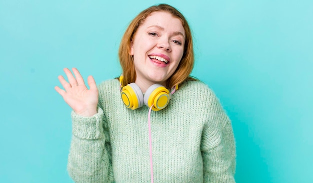 Hübsche Frau mit rotem Kopf, die glücklich lächelt und mit der Hand winkt, die Sie begrüßt und grüßt, wenn Sie das Musikkonzept genießen