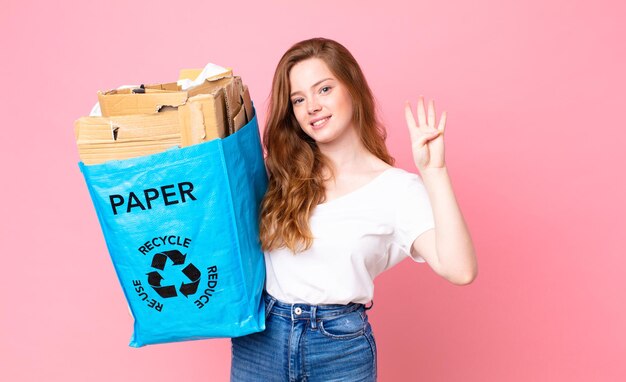 Hübsche Frau mit rotem Kopf, die lächelt und freundlich aussieht, Nummer vier zeigt und eine Recyclingpapiertüte hält