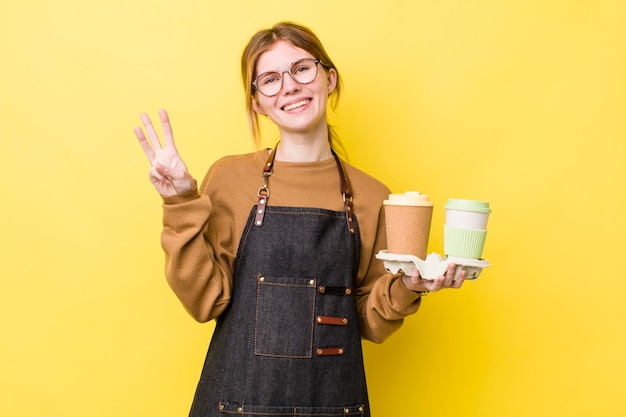 Hübsche Frau mit rotem Kopf, die lächelt und freundlich aussieht und das Konzept Nummer drei zum Mitnehmen von Kaffee zeigt