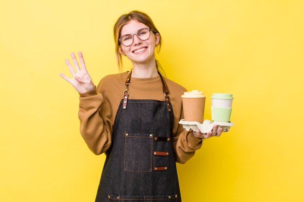 Hübsche Frau mit rotem Kopf, die lächelt und freundlich aussieht und das Konzept Nummer vier zum Mitnehmen von Kaffee zeigt