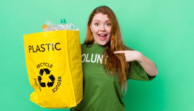 Hübsche Frau mit rotem Kopf, die sich glücklich fühlt und mit einem aufgeregten Recyclingkonzept auf sich selbst zeigt