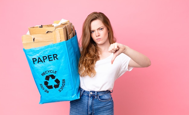 Hübsche Frau mit rotem Kopf, die sich überquert, Daumen nach unten zeigt und eine Recyclingpapiertüte hält