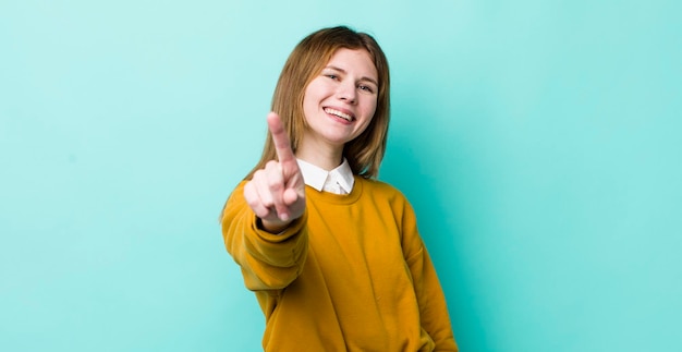 Hübsche Frau mit rotem Kopf, die stolz und selbstbewusst lächelt und die Pose Nummer eins triumphierend macht und sich wie ein Anführer fühlt