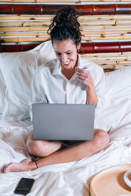 Hübsche Frau mit Telefon und Laptop im Bett