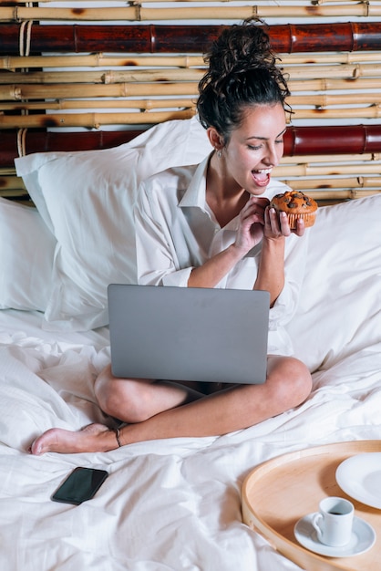 Hübsche Frau mit Telefon und Laptop im Bett