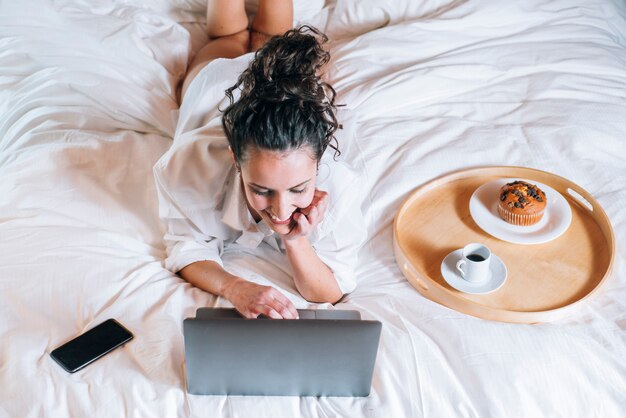 Hübsche Frau mit Telefon und Laptop im Bett